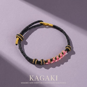 May include: Black and gold braided bracelet with pink accents. The bracelet features a knot closure and decorative gold beads. The word "KAGAKI" is printed at the bottom of the image, along with the phrase "DESIGNED WITH INTENTION • HANDMADE WITH BLESSINGS."