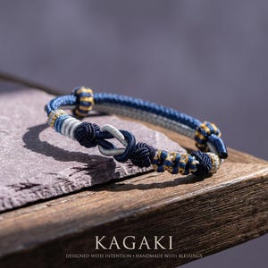 Puede incluir: Una pulsera artesanal con intrincados nudos en tonos azul, gris y dorado. La pulsera presenta un diseño de nudo central y se exhibe sobre una superficie de madera. El texto "KAGAKI" es visible.