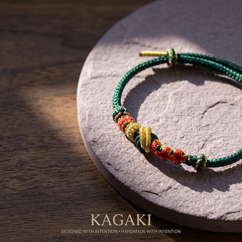 May include: A handcrafted green and gold bracelet with red and gold accents. The bracelet is made of woven cord and features intricate knotting. The word "KAGAKI" is displayed at the bottom of the image, along with the text "DESIGNED WITH INTENTION &bull; HANDMADE WITH INTENTION."