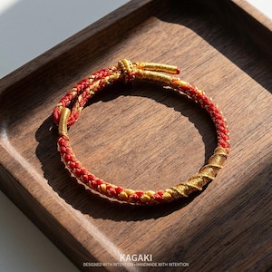 May include: Red and gold braided bracelet with a knotted closure. The bracelet is displayed on a wooden tray. The text "KAGAKI DESIGNED WITH INTENTION HANDMADE WITH INTENTION" is at the bottom of the image.