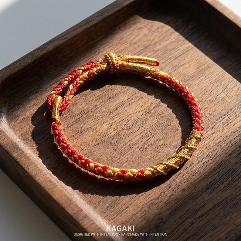 May include: Red and gold braided bracelet with a knotted closure. The bracelet is displayed on a wooden tray. The text "KAGAKI DESIGNED WITH INTENTION HANDMADE WITH INTENTION" is at the bottom of the image.