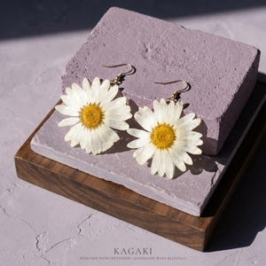 Op de afbeelding: Een paar madeliefjesoorbellen met goudkleurige haken. Elke oorbel heeft een witte madeliefbloem met een gele kern. De oorbellen worden getoond op een paars blok en een houten dienblad. De tekst "KAGAKI" staat onderaan.