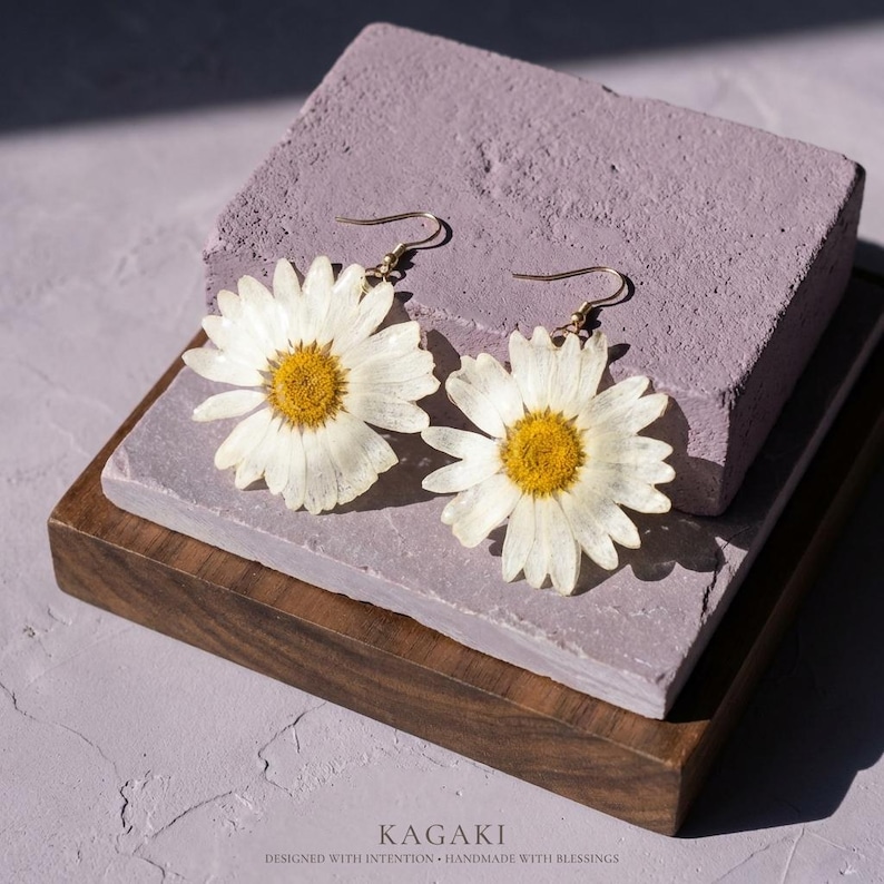 Op de afbeelding: Een paar madeliefjesoorbellen met goudkleurige haken. Elke oorbel heeft een witte madeliefbloem met een gele kern. De oorbellen worden getoond op een paars blok en een houten dienblad. De tekst "KAGAKI" staat onderaan.