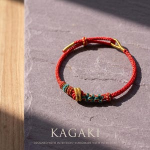 May include: Red braided bracelet with gold-colored accents and decorative beads in teal, red, and gold. The bracelet is on a gray surface. The word "KAGAKI" is displayed below the bracelet.