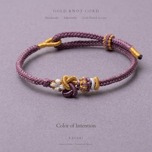 Puede incluir: Una pulsera de cordón morado ajustable con detalles chapados en oro y un nudo central. El texto dice GOLD KNOT CORD, Handmade, Adjustable, Gold-Plated Accent, Color of Intention, KAGAKI, Inspired by Nature, Handmade with Intention.