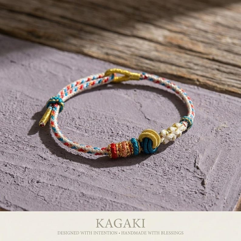 May include: A handmade bracelet with a woven design in white, red, blue, and gold. The bracelet features a knot design with colorful beads and the text "KAGAKI" below. The bracelet is designed with intention and handmade with blessings.