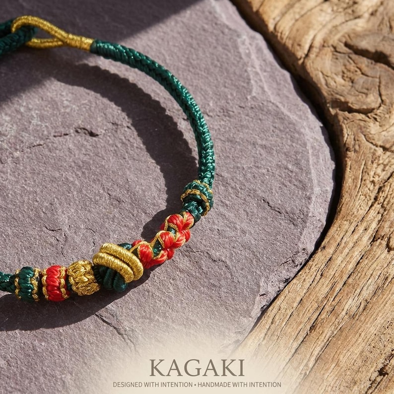 May include: A handcrafted green, red, and gold bracelet. The bracelet features intricate knotting and braided details. The word "KAGAKI" is printed at the bottom of the image, along with the text "DESIGNED WITH INTENTION &bull; HANDMADE WITH INTENTION."