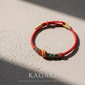 May include: Red braided bracelet with gold-colored accents and decorative knots in teal and yellow. The bracelet is handmade and features the text "KAGAKI" and "DESIGNED WITH INTENTION &bull; HANDMADE WITH INTENTION".