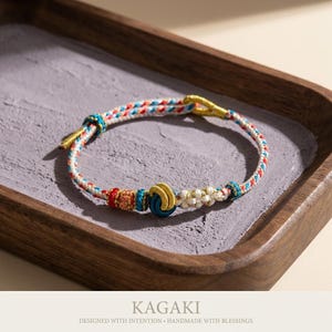 May include: A handmade bracelet with a woven cord in white, red, and blue. The bracelet features decorative knots and gold-colored metal accents. The text "KAGAKI" and "DESIGNED WITH INTENTION &bull; HANDMADE WITH BLESSINGS" are at the bottom.