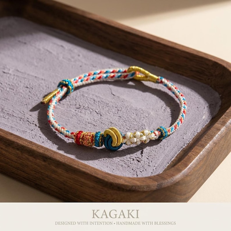 May include: A handmade bracelet with a woven cord in white, red, and blue. The bracelet features decorative knots and gold-colored metal accents. The text "KAGAKI" and "DESIGNED WITH INTENTION &bull; HANDMADE WITH BLESSINGS" are at the bottom.