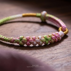 Pode incluir: Uma pulseira trançada com um padrão repetitivo de cordões rosa, branco e verde claro entrelaçados com fio dourado metálico. O design inclui uma seção central de nós decorativos e um anel conector dourado. O texto diz KAGAKI.