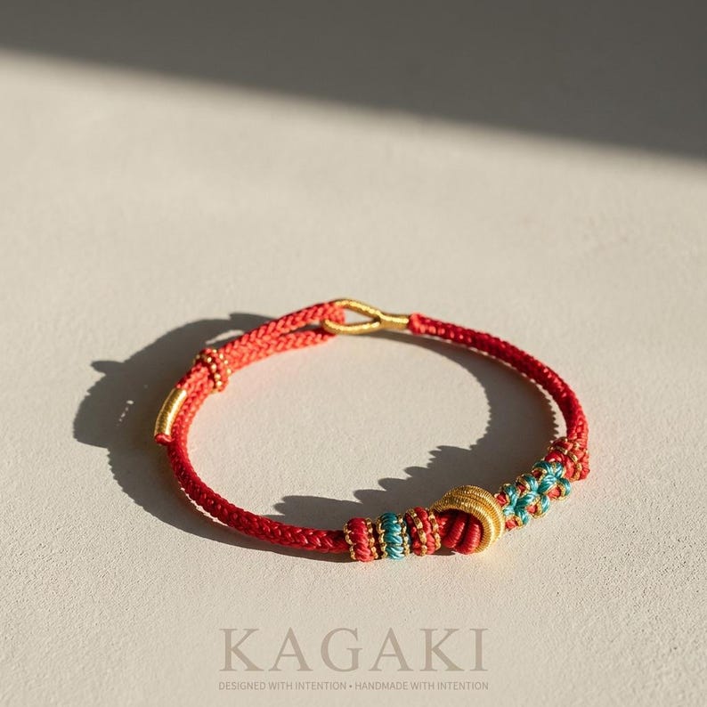 May include: Red braided bracelet with gold accents and turquoise and gold detailing. The bracelet features the word "KAGAKI" and the text "DESIGNED WITH INTENTION + HANDMADE WITH INTENTION".