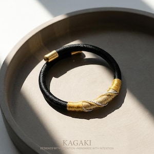 May include: Black bracelet with gold-colored accents. The bracelet features a black band with a gold clasp and a section wrapped in gold and silver-colored threads. The bracelet is displayed on a gray tray.