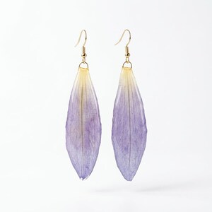 May include: A pair of handmade earrings featuring delicate, elongated petals in shades of purple and yellow. The petals are suspended from gold-colored ear wires, creating a unique and natural accessory. The earrings are set against a clean white background.