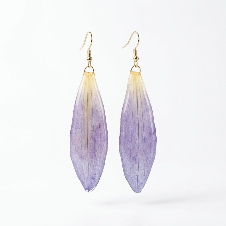 May include: A pair of handmade earrings featuring delicate, elongated petals in shades of purple and yellow. The petals are suspended from gold-colored ear wires, creating a unique and natural accessory. The earrings are set against a clean white background.