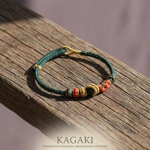 May include: A handmade green bracelet with gold accents and red and gold decorative knots. The bracelet is displayed on a wooden surface. The text "KAGAKI" and "DESIGNED WITH INTENTION - HANDMADE WITH INTENTION" are at the bottom.