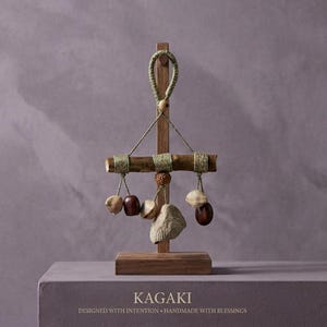 May include: A wooden decorative mobile with a rectangular base and a vertical support. A horizontal wooden bar is suspended from the top, adorned with hanging natural elements in various shapes and colors. The text "KAGAKI" is at the bottom.