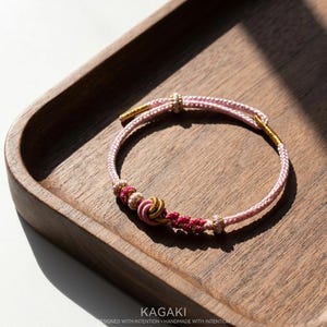 May include: A pink and gold bracelet with intricate knotting and beadwork. The bracelet is set on a wooden tray. The text "KAGAKI" is visible at the bottom of the image, along with the phrase "DESIGNED WITH INTENTION - HANDMADE WITH INTENTION."