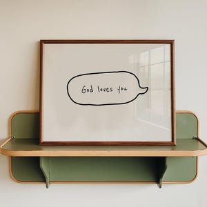 May include: A framed print with the text "God loves you" in a speech bubble. The print is displayed on a two-tiered shelf with a green base and a wooden frame. The shelf is mounted on a white wall.