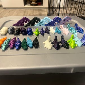 May include: A selection of brightly coloured, 3D-printed dinosaur and lizard figurines. The collection features articulated designs in shades of purple, black, blue, green, and orange. The figurines are displayed on a grey plastic storage container.