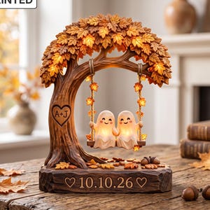Puede incluir: Un adorno decorativo plano en 2D que muestra dos fantasmas sonrientes sentados en un columpio suspendido de un árbol de otoño con hojas naranjas. El tronco tiene un corazón con las letras A+J y la base muestra la fecha 10.10.26.