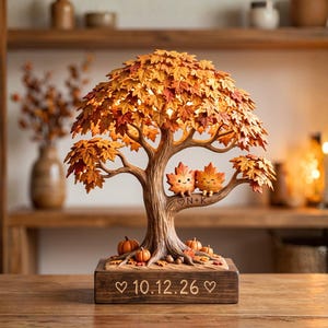 Puede incluir: Una escultura decorativa de árbol de otoño con hojas de arce naranjas y amarillas, con dos personajes de hoja sonrientes en una rama grabada con un corazón y N+K. La base de madera rectangular está grabada con la fecha 10.12.26 y dos corazones.