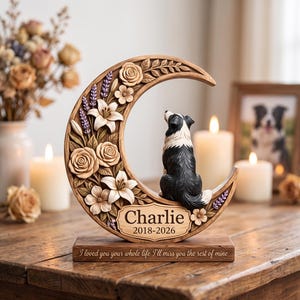 Puede incluir: Una placa conmemorativa de madera en forma de media luna con rosas y lirios tallados, y una figura de perro blanco y negro sentada en la curva. La placa muestra el nombre Charlie, los años 2018-2026 y el texto I loved you your whole life.