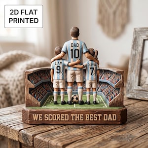 May include: A 2D flat printed figurine of a soccer player with the name DAD and number 10, standing with two children named LIAM with number 9 and OLIVIA with number 7. They wear light blue and white jerseys on a field with the text WE SCORED THE BEST DAD.