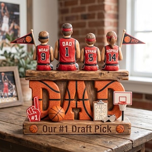 May include: A basketball-themed decorative figurine featuring a family of four, with the word "DAD" prominently displayed. The figures wear red jerseys with numbers and hold flags. Additional elements include a basketball, a coach's clipboard, and the text "Our #1 Draft Pick."