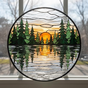 Könnte beinhalten: Runde Glasmalerei mit einem Sonnenuntergang über einem See mit immergrünen Bäumen. Die Sonne ist leuchtend orange und die Bäume sind grün. Der See spiegelt den Sonnenuntergang und die Bäume wider. Das Panel ist schwarz gerahmt und hängt an einer Kette.