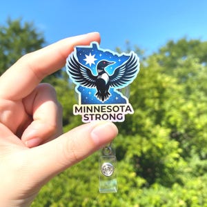 May include: A badge reel featuring a design of the state of Minnesota with a loon and the text "MINNESOTA STRONG". The badge reel is held in a hand against a backdrop of green foliage and a blue sky.