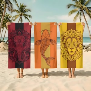 May include: Three beach towels with unique designs are held up on a sandy beach. One towel features a red elephant design, another a koi fish, and the last a lion's head. Each towel has a vertical stripe design.