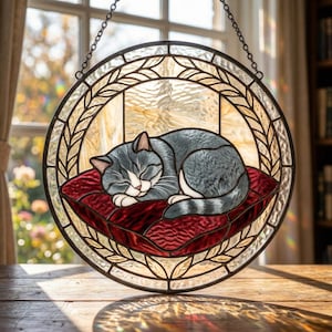 May include: Round stained glass window art featuring a sleeping gray and white cat on a red pillow. The cat is curled up, eyes closed, within a circular frame with a leaf design. The window is suspended by a chain.