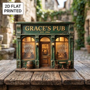 Op de afbeelding: Een miniatuurmodel van een Ierse pub-gevel met een verweerde groene afwerking, voorzien van de tekst "GRACE'S PUB EST. 1986" boven de ingang. De gevel bevat twee ramen met "IRISH PUB"-stickers, een centrale deur, twee kleine lantaarns en een vat.