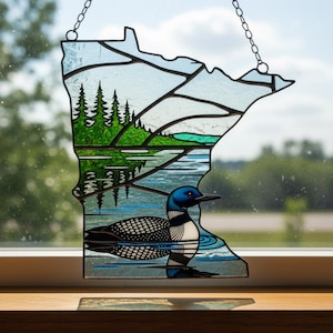 May include: A stained glass suncatcher shaped like the state of Minnesota, featuring a black and white loon swimming on blue water, a green forest of pine trees, and a light blue sky, all held together by dark metal solder lines and a hanging chain.