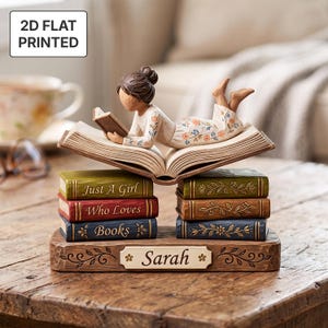 Puede incluir: Una figura decorativa de una persona acostada sobre un libro abierto, que descansa sobre dos pilas de tres libros. Los libros son verdes, rojos y azules, con el texto "Just A Girl Who Loves Books" y una placa base "Sarah" con detalles florales.