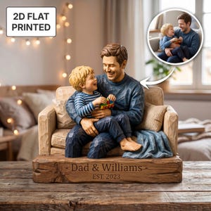 May include: A 2D flat printed figurine of a father and child sitting on a couch, with the child holding a toy train. The base is engraved with the text 'Dad & Williams - EST. 2023'. The figures have a carved wood texture and blue clothing.