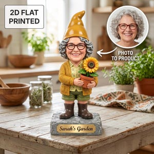 Op de afbeelding: Een grillige tuinkabouterfiguur met een lachend gezicht, een bril, een gele hoed en een vest. De kabouter houdt een zonnebloem in een pot vast. De basis luidt "Sarah's Garden". De afbeelding bevat ook de tekst "2D FLAT PRINTED" en "PHOTO TO PRODUCT".