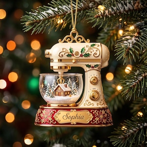 Könnte beinhalten: Ein cremefarbener Weihnachtsbaumschmuck in Form einer Küchenmaschine mit goldenen Akzenten, Stechpalmenmotiven und einer Glasschüssel, die eine Miniatur-Blockhütte und Tannenbäume enthält. Der rote Sockel zeigt goldene Blumenmuster und den Namen Sophia.