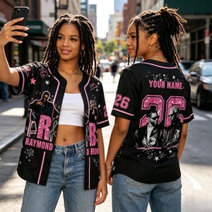 May include: A black baseball jersey with pink glitter accents, featuring the text "R&B", "RAYMOND", "YOUR NAME", and the number "26". The design includes graphic prints of two men in sunglasses, white star motifs, and pink trim on the sleeves and collar.