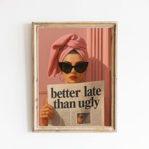 May include: Framed art print featuring a woman wearing a pink towel turban and black sunglasses, holding a newspaper with the headline "better late than ugly." The artwork has a vintage aesthetic with a pink and beige color palette.