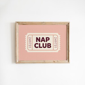 Può includere: Una stampa incorniciata con sfondo rosa e design a biglietto. Il biglietto recita "NAP CLUB" in lettere marrone scuro. La cornice è di colore marrone chiaro. La stampa è un oggetto decorativo.