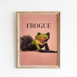 Op de afbeelding: Ingelijste kunstdruk met een groene kikker in een zwarte jurk, handschoenen en een parelarmband, met rode lippenstift en lange wimpers. Het woord "FROGUE" staat in zwarte letters bovenaan op een roze achtergrond.