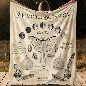 May include: A cream-colored throw blanket featuring botanical illustrations and text. The design includes a large moth, phases of the moon, leaves, inkwells, and feathers. Text includes "Grimoire Botanica", "Luna Moth", "Amor Fati", and "Ad Astra Per Aspera".