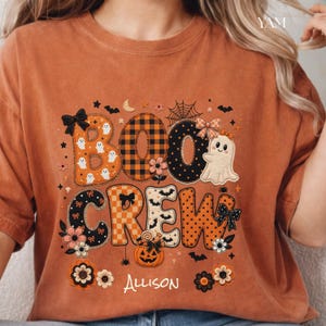 May include: A rust-colored t-shirt featuring a graphic with the text "BOO CREW" and "Allison". The letters are decorated with patterns including plaid, polka dots, ghosts, bats, bows, flowers, a spider web, and a jack-o'-lantern.