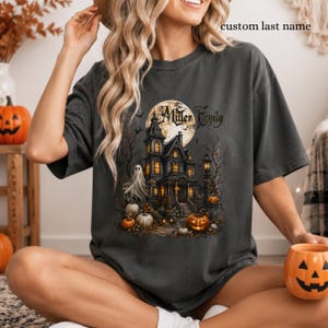 Puede incluir: Una camiseta gris carbón con un estampado gráfico de una mansión encantada bajo una luna llena, rodeada de calabazas, un fantasma y árboles desnudos. El texto "The Miller Family" está impreso encima, con "custom last name" arriba.