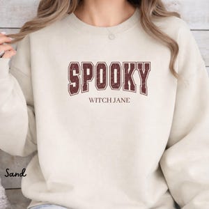 Puede incluir: Una sudadera de cuello redondo color arena que presenta la palabra SPOOKY en una fuente universitaria grande de color marrón oscuro con efecto desgastado, con el texto WITCH JANE impreso en una fuente serif marrón simple debajo.