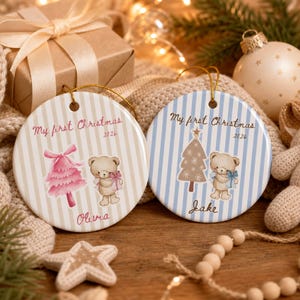 Puede incluir: Dos adornos navideños redondos de cerámica con cordones dorados. Uno tiene rayas beige, un árbol rosa, un oso de peluche y el texto My first Christmas 2026 Olivia. El otro tiene rayas azules, un árbol, un oso y el texto My first Christmas 2026 Jake.