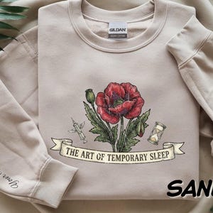 May include: A sand-colored crewneck sweatshirt featuring a central graphic of a red poppy flower, a syringe, and an hourglass. A banner below the illustration displays the text THE ART OF TEMPORARY SLEEP. The sleeve includes the text YOUR NAME.