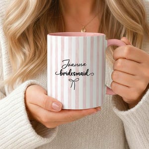 Puede incluir: Una taza a rayas blancas y rosas con asa e interior rosas. La taza tiene las palabras "Joanna bridesmaid" en escritura negra con un lazo gráfico. La taza está sostenida por una persona.